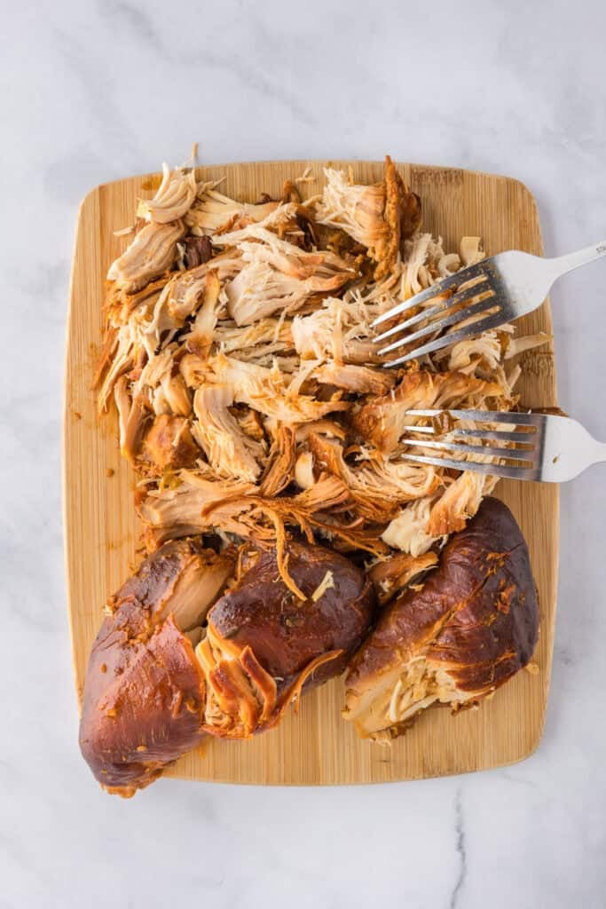 Cooked chicken being shredded on a wooden cutting board with two forks for Slow Cooker BBQ Hawaiian Roll Chicken Sliders.