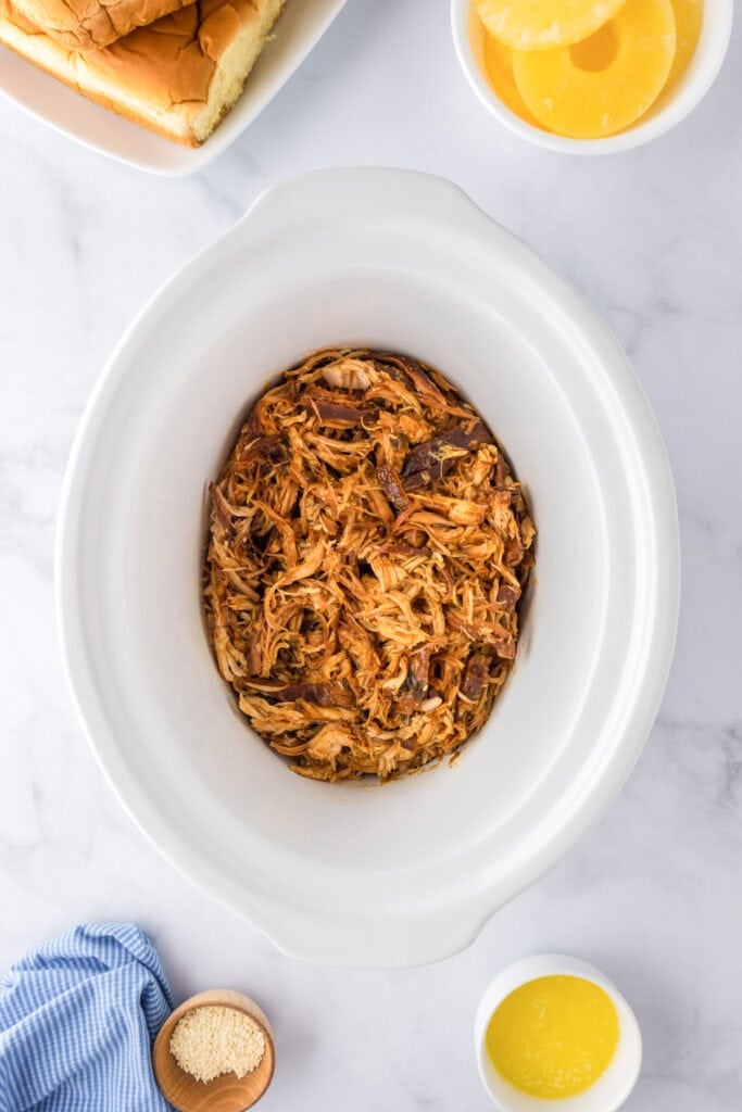 Shredded chicken in a slow cooker, surrounded by bowls of pineapple slices, melted butter, sesame seeds, and a plate of sandwich buns ready to assemble Slow Cooker BBQ Hawaiian Roll Chicken Sliders on a marble countertop.