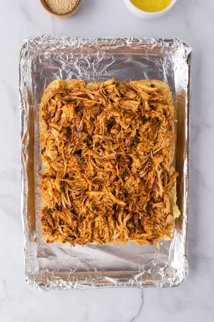 A sheet of Slow Cooker BBQ Hawaiian Roll Chicken Sliders open face on a foil-lined baking tray being prepared.