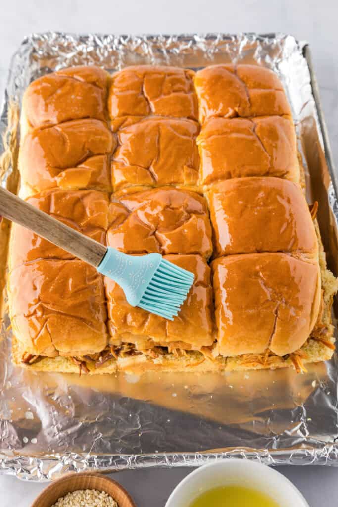 A brush spreads melted butter over a tray of Slow Cooker BBQ Hawaiian Roll Chicken Sliders on foil, with bowls of sesame seeds and extra melted butter nearby.