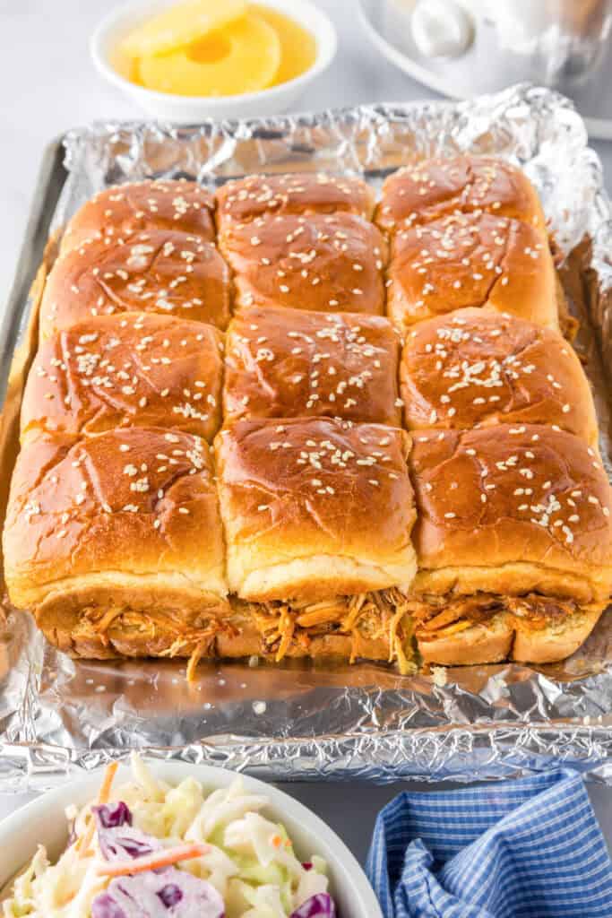 A baking tray lined with foil holds twelve Slow Cooker BBQ Hawaiian Roll Chicken Sliders topped with sesame seeds; shredded chicken filling is visible. Coleslaw and a bowl of pineapple slices are in the background.