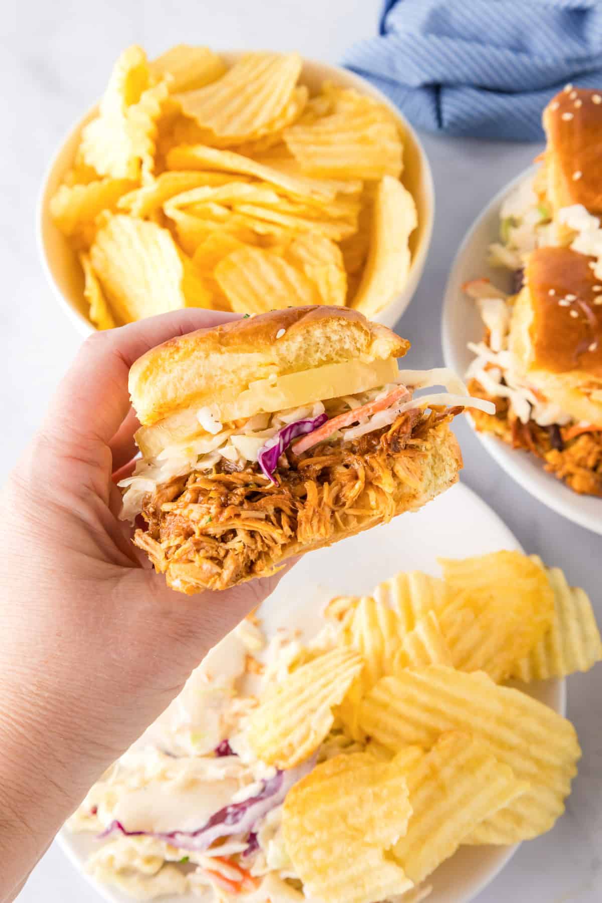 A hand holds a Slow Cooker BBQ Hawaiian Roll Chicken Slider topped with coleslaw. Plates and bowls of potato chips and more sliders are visible in the background.