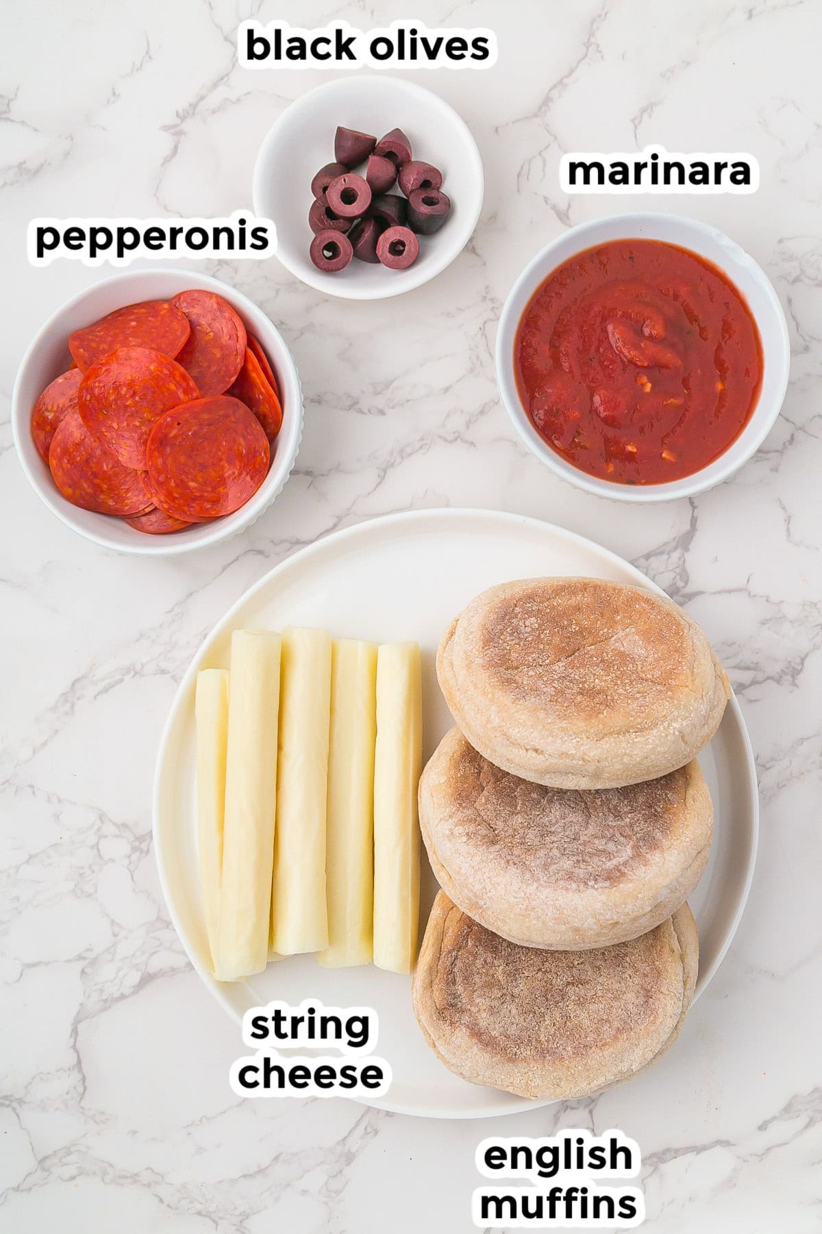 A plate with three English muffins and string cheese sticks, next to bowls of pepperoni slices, black olives, and marinara sauce, all labeled for easy mummy pizzas.
