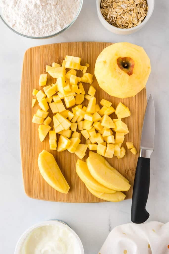 A peeled apple is diced and sliced on a wooden cutting board with a paring knife, surrounded by bowls of flour, oats, and yogurt—ingredients ready for making an apple dessert pizza.