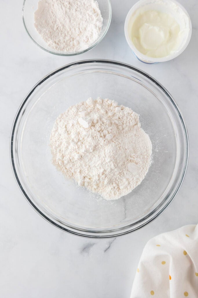 A glass mixing bowl containing flour, ready for making apple dessert pizza, sits on a white surface with extra bowls of flour and a bowl of yogurt nearby.