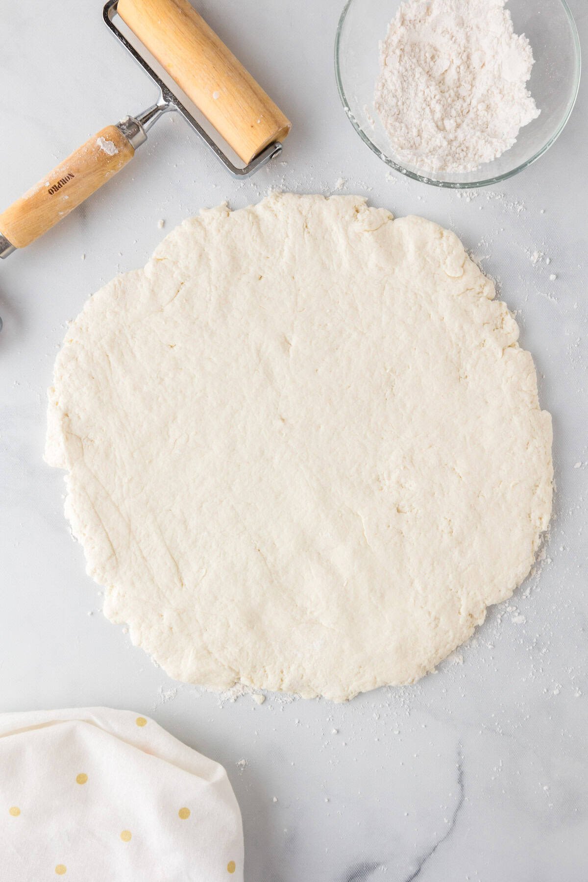 Rolled out dough on a floured surface for an apple dessert pizza, with a rolling pin, a bowl of flour, and a polka dot cloth nearby.
