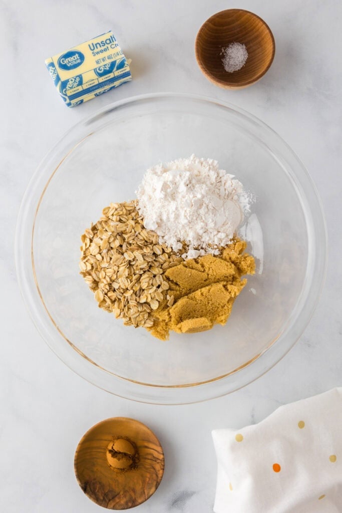 A glass bowl with oats, powdered sugar, and brown sugar for making the crumble on top of an apple dessert pizza. Nearby are a stick of butter, bowls of salt and cinnamon, and a dotted white cloth.