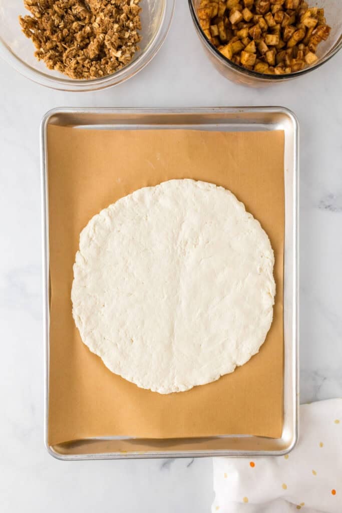 A round, uncooked apple dessert pizza dough rests on a parchment-lined baking sheet, with bowls of sweet toppings nearby on a marble surface.
