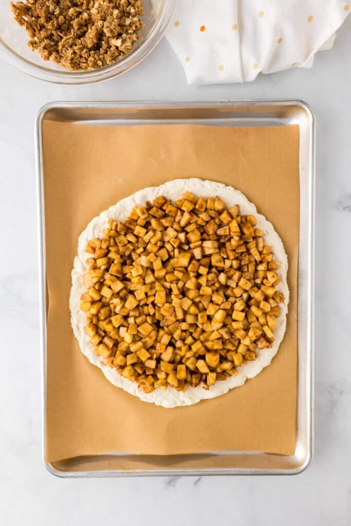 Unbaked pizza on a parchment-lined baking sheet, topped with chopped cinnamon apples for an apple dessert pizza. A bowl with crumble topping is partially visible above.