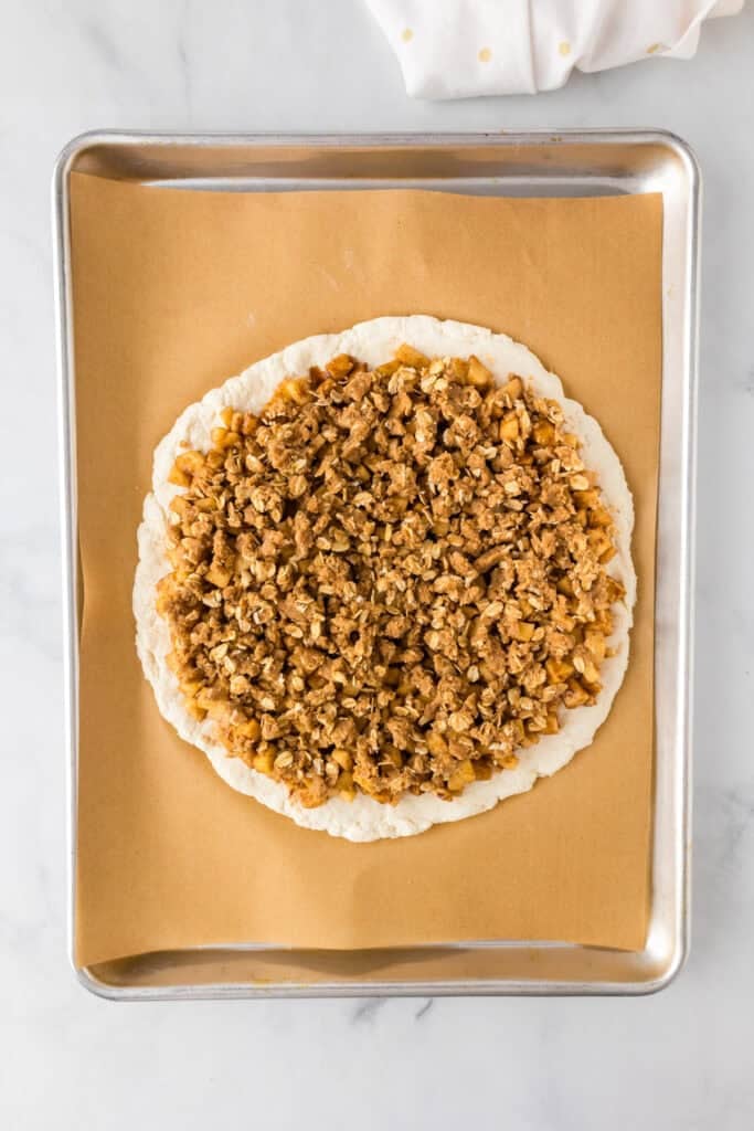 Unbaked round dough topped with a mixture of chopped apples and crumble topping on a parchment-lined baking sheet, ready to become a delicious apple dessert pizza.