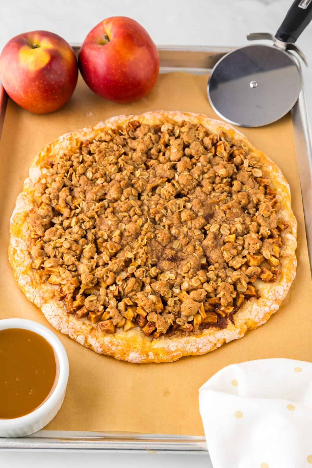 A baked apple dessert pizza sits on parchment paper beside two apples, a pizza cutter, a small bowl of caramel sauce, and a white napkin with yellow dots.