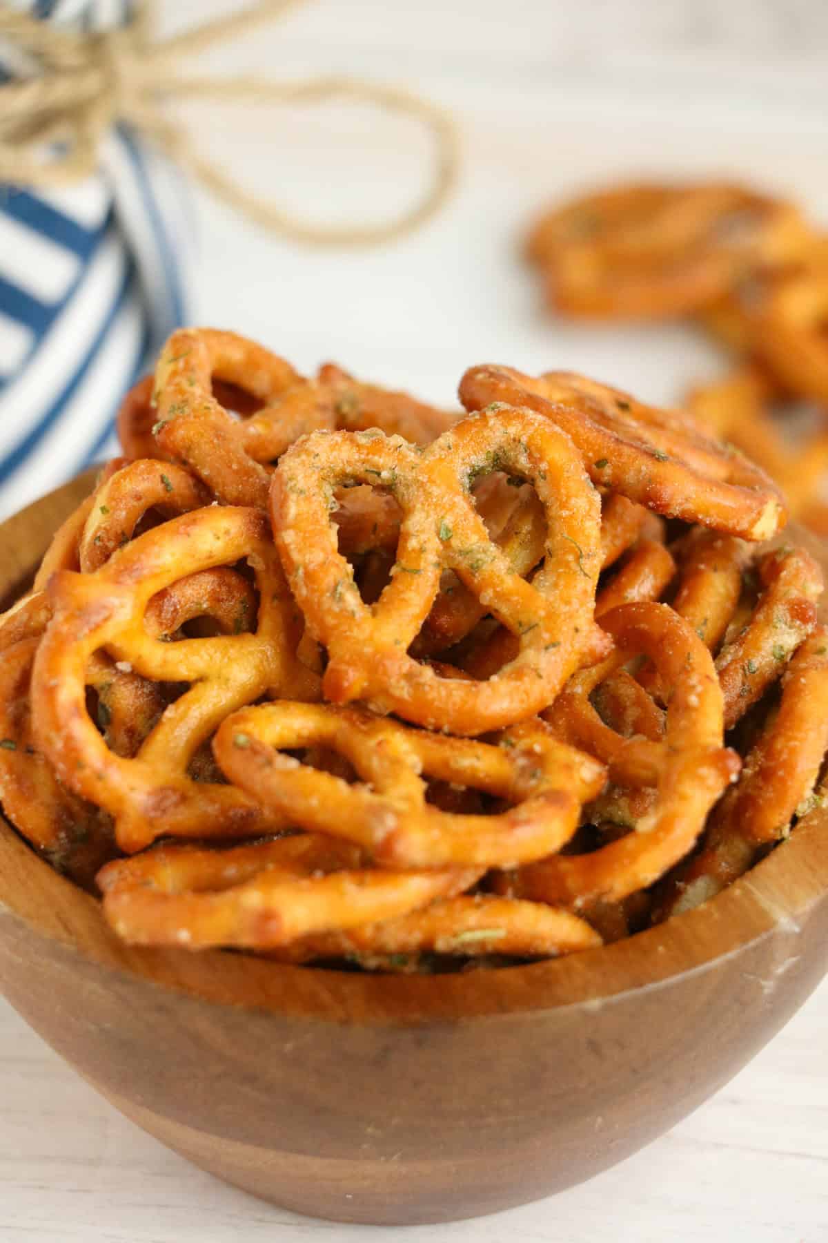 A wooden bowl filled with garlic ranch pretzels sits on a light surface, with more pretzels and a blue-striped cloth in the background.