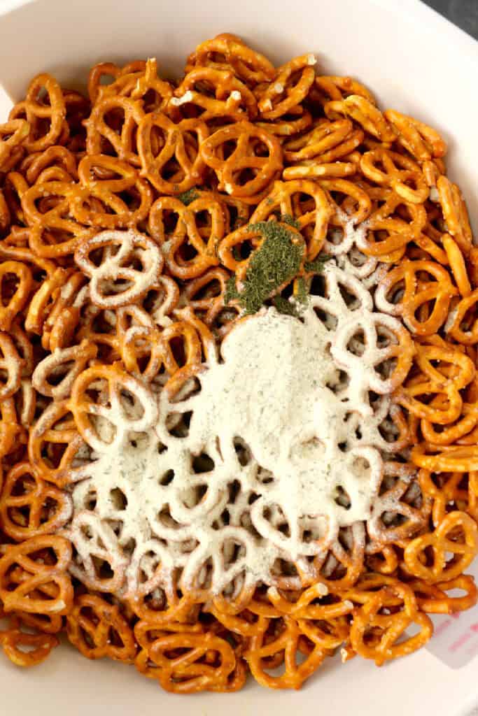 A bowl of mini garlic ranch pretzels topped with powdered ranch seasoning and dried herbs, ready for mixing.
