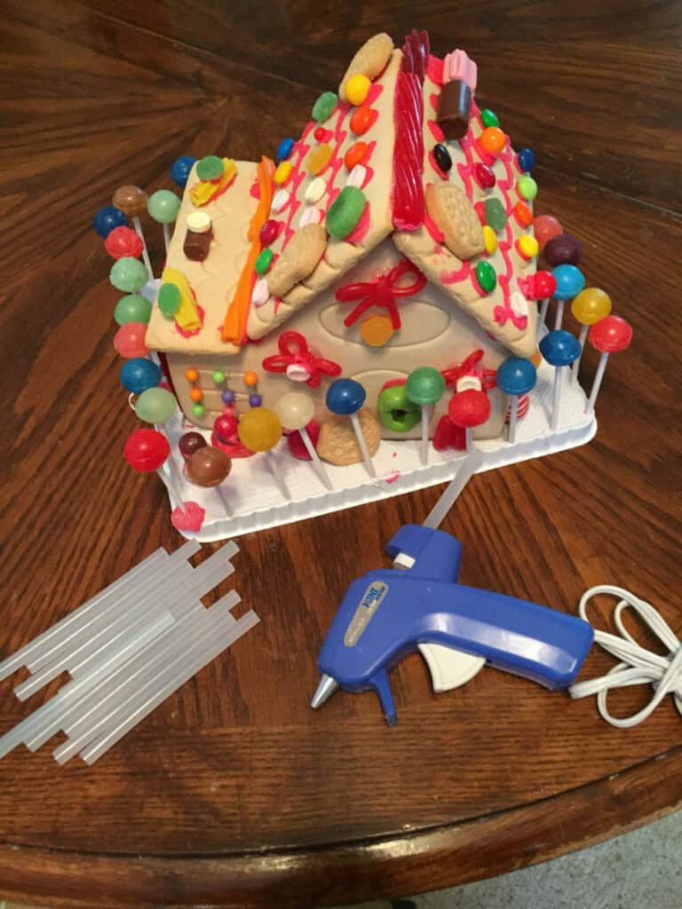 A decorated candy-covered gingerbread house on a table with a blue hot glue gun and glue sticks beside it.
