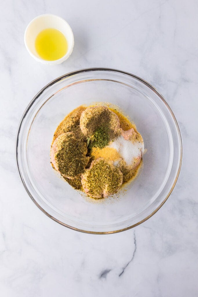 A glass bowl with raw ground turkey meatballs mixture and various spices sits on a white marble surface. A small bowl with oil is placed nearby.