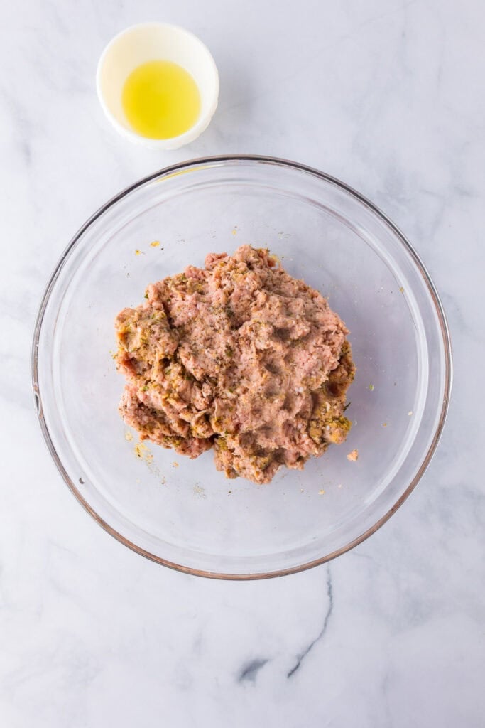 A glass bowl containing a raw ground turkey meatball mixture sits on a marble surface next to a small bowl of oil.