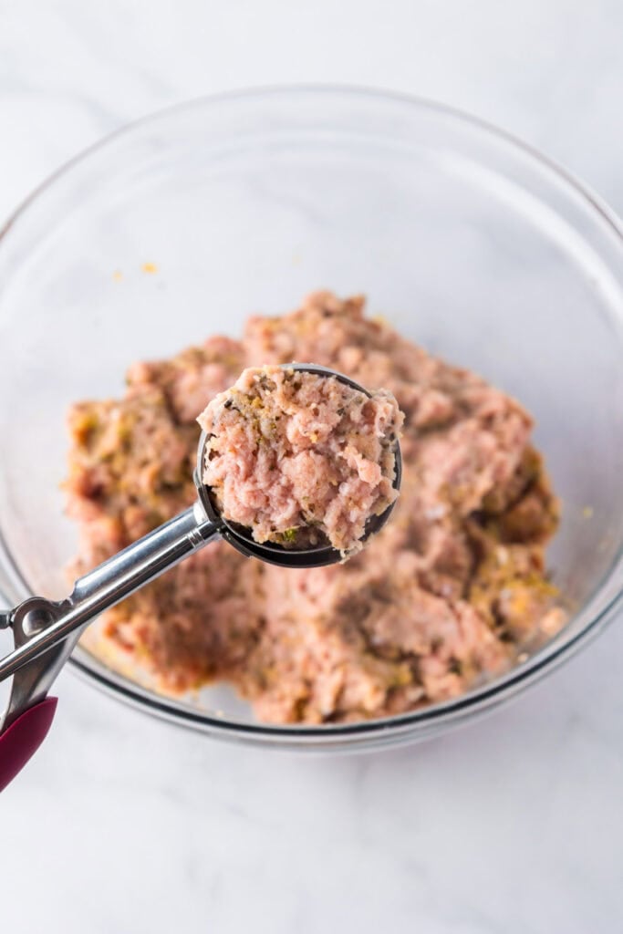 A metal scoop holds a portion of raw ground turkey meatballs mixture above a glass bowl filled with more of the same mixture.