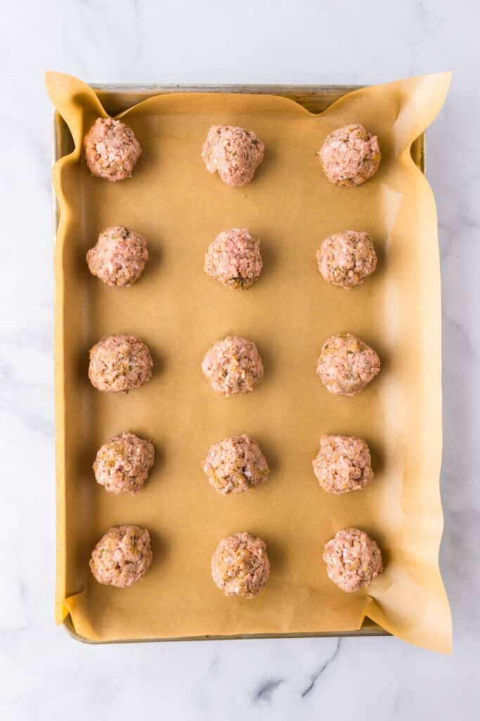 A baking sheet lined with parchment paper holds fifteen evenly spaced raw ground turkey meatballs, ready to be cooked.