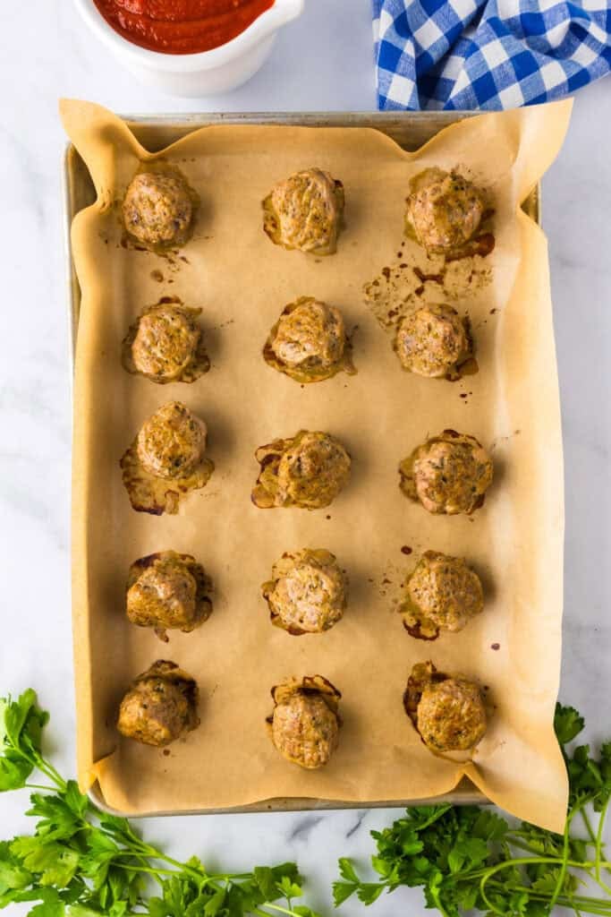 A baking sheet lined with parchment paper holds fifteen cooked ground turkey meatballs, with a bowl of tomato sauce, a blue checkered cloth, and parsley nearby.