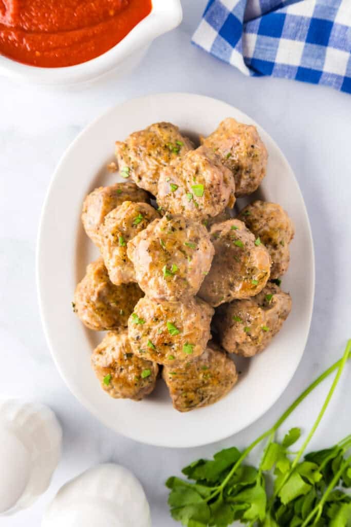 A white oval plate with a stack of cooked ground turkey meatballs next to a bowl of red sauce on a table.