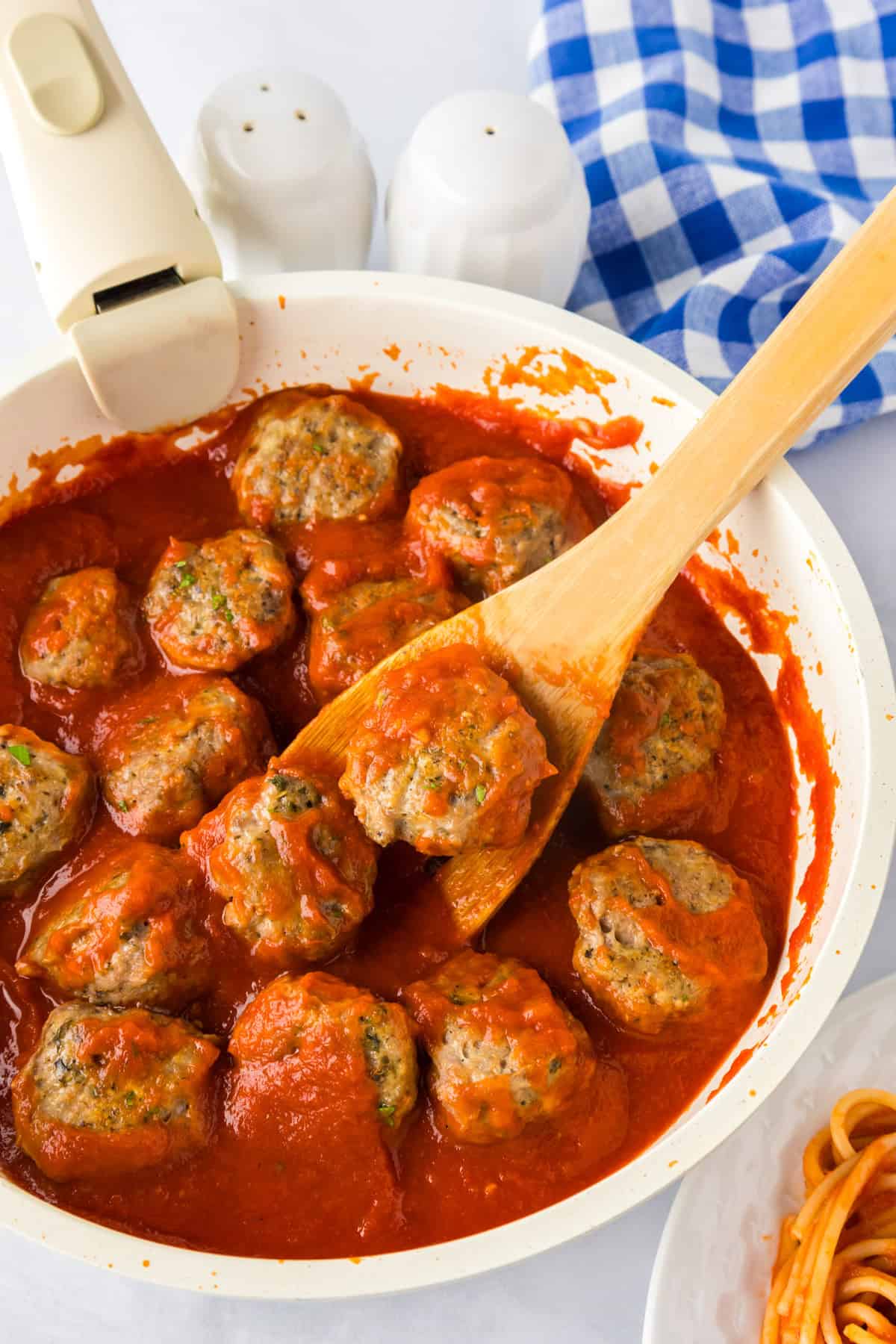 A white skillet filled with ground turkey meatballs in tomato sauce; a wooden spoon lifts one meatball. A plate of spaghetti and salt and pepper shakers are nearby.