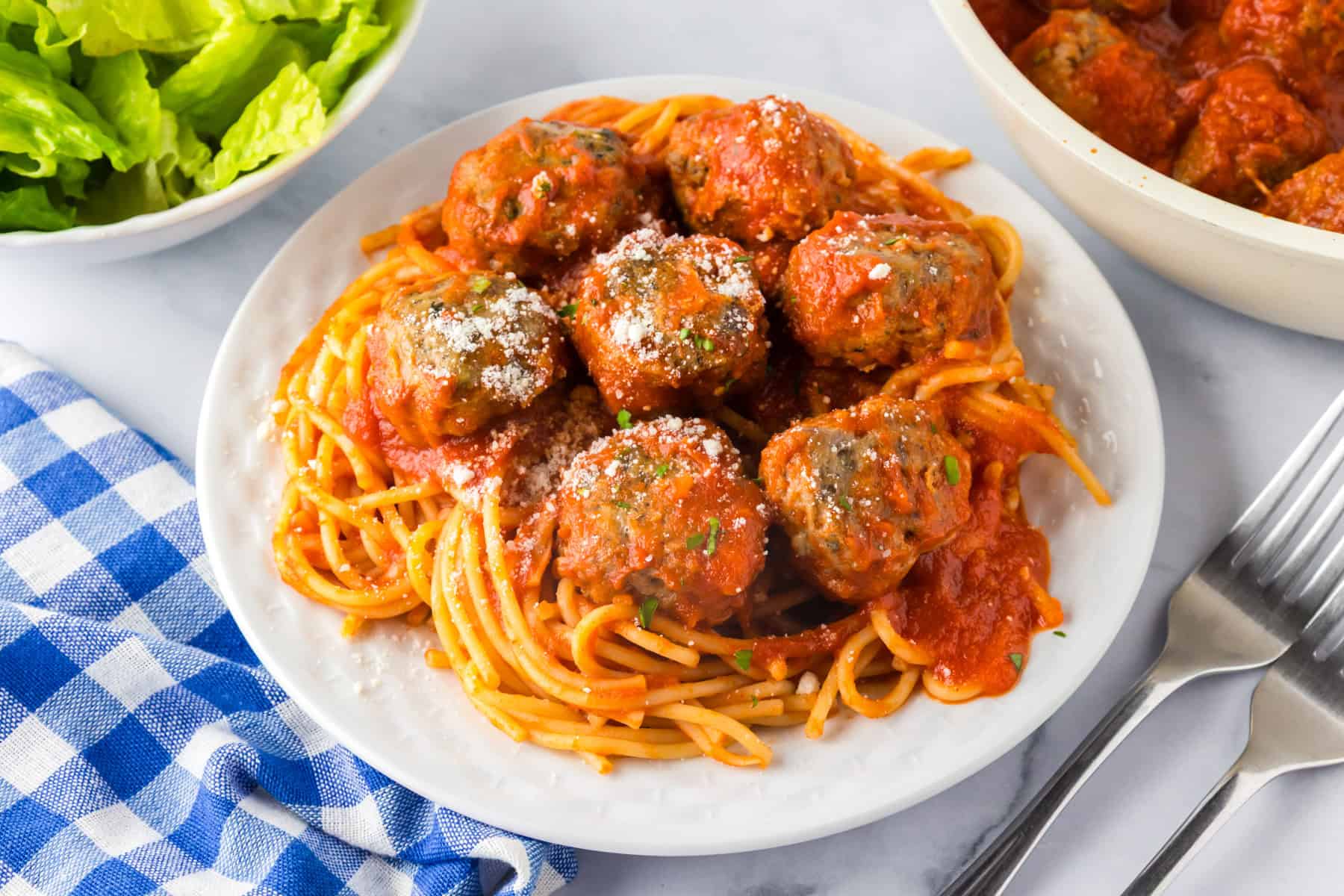 A plate of spaghetti topped with tomato sauce and several ground turkey meatballs, garnished with grated cheese, sits beside a bowl of lettuce and a blue checkered napkin.