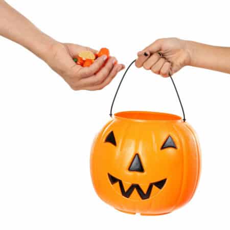 A person dropping Halloween candies into a plastic pumpkin bucket held by another person, against a white background.