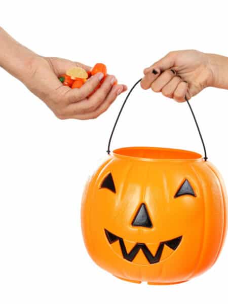 A person dropping Halloween candies into a plastic pumpkin bucket held by another person, against a white background.