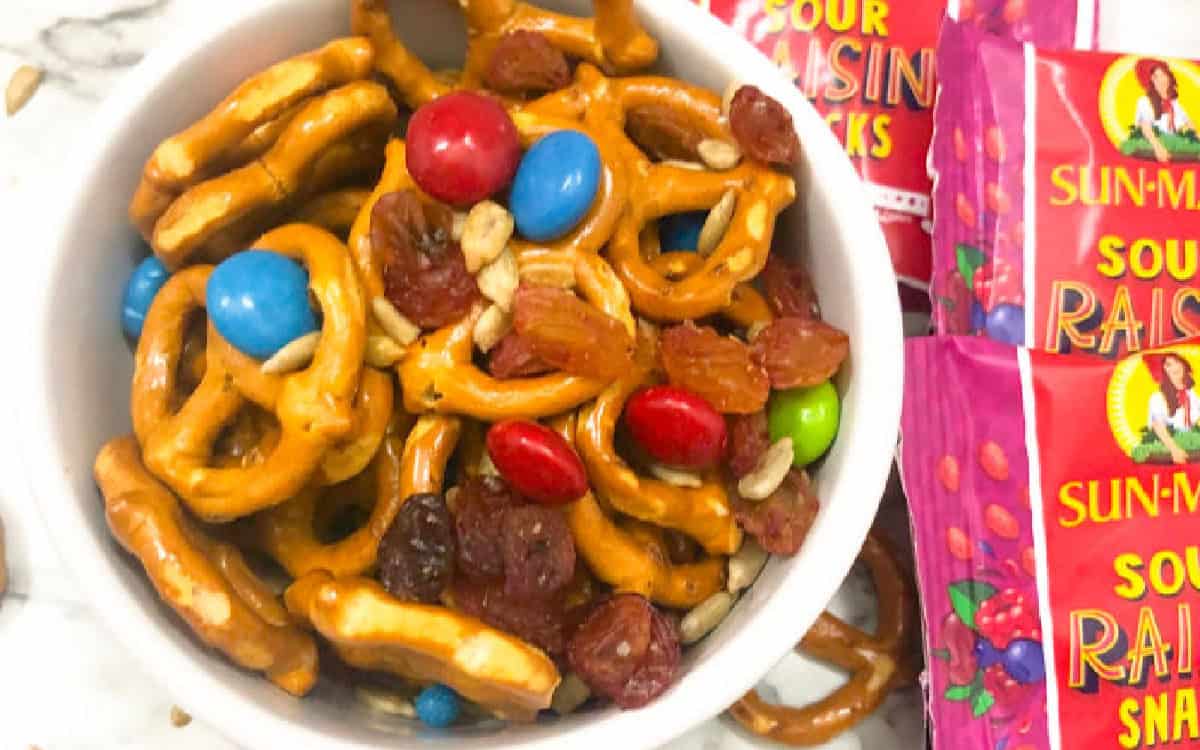 A white bowl filled with trail mix including pretzels, colorful candy-coated chocolates, raisins, and sunflower seeds.