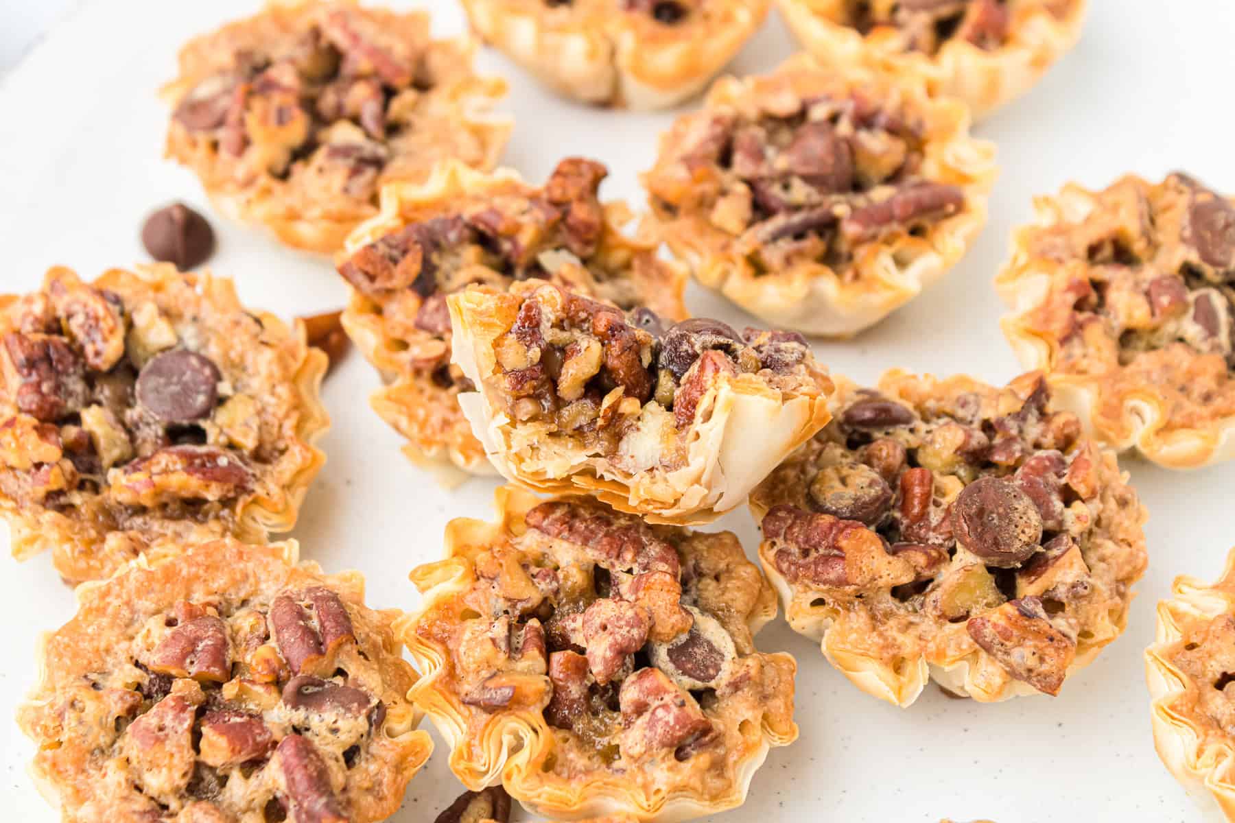 Mini pecan pie bites in phyllo cups with chocolate chips arranged on a white surface, bite size pecan tartlets, one cut open to show gooey filling.