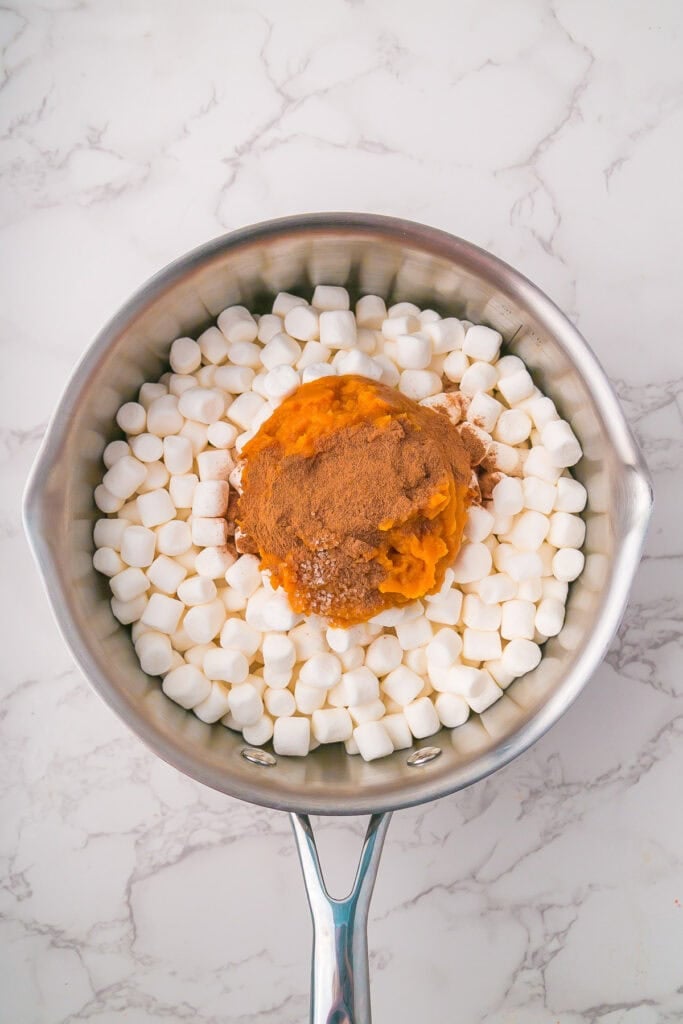 A saucepan on a marble surface contains mini marshmallows, pumpkin puree, and ground spices arranged in separate piles, ready to be mixed for a delicious pumpkin marshmallow pie.