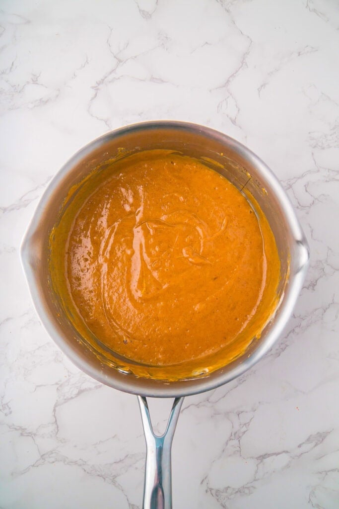 A stainless steel saucepan filled with a smooth, orange-brown pumpkin marshmallow pie mixture sits on a white marble surface.
