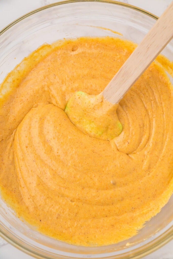 A glass bowl filled with smooth, orange pumpkin marshmallow pie batter is being stirred with a wooden spatula after folding in whipped topping.