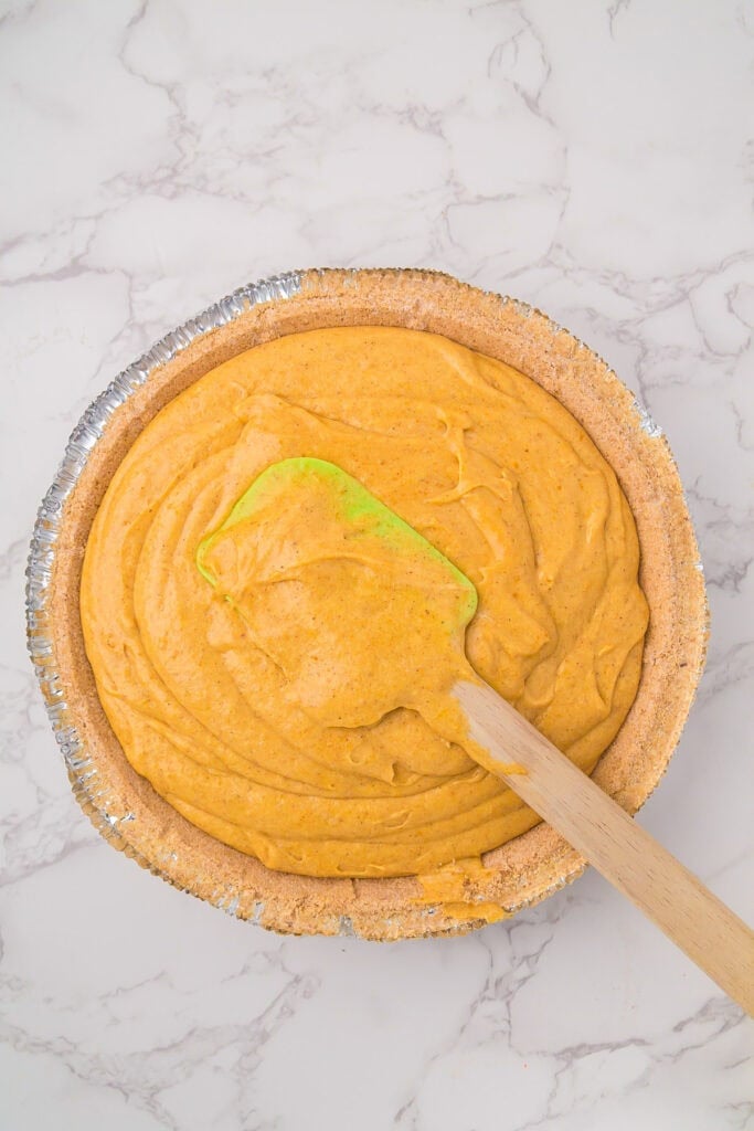 A pie crust filled with creamy orange pumpkin marshmallow pie filling is being smoothed with a green and wooden spatula on a marble surface.