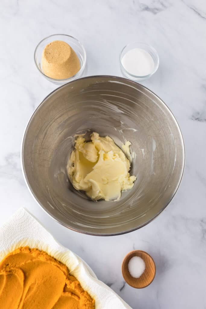 A mixing bowl with creamed butter, surrounded by bowls of brown sugar, white sugar, salt, and a plate with pumpkin puree on a marble surface to make Pumpkin Chocolate Chip Bars.