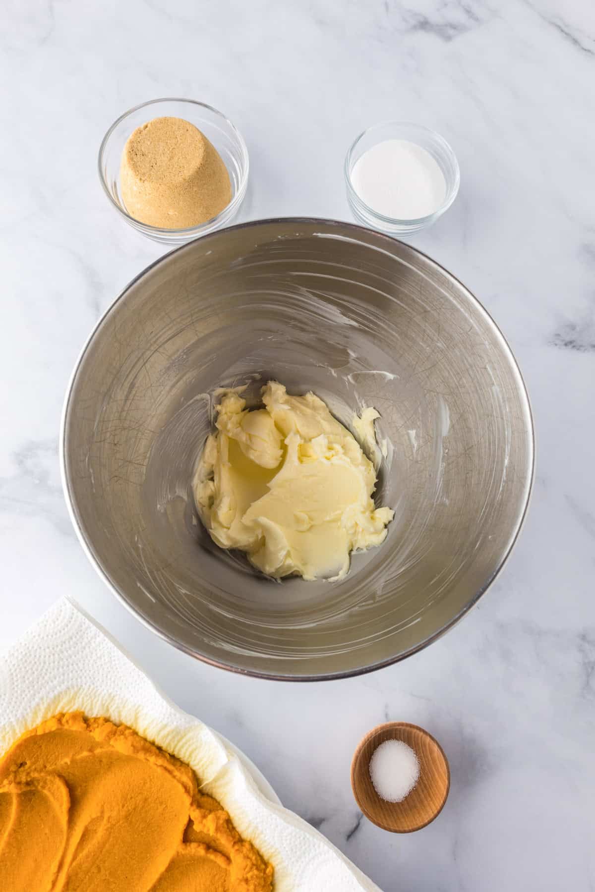 A mixing bowl with creamed butter, surrounded by bowls of brown sugar, white sugar, salt, and a plate with pumpkin puree on a marble surface to make Pumpkin Chocolate Chip Bars.