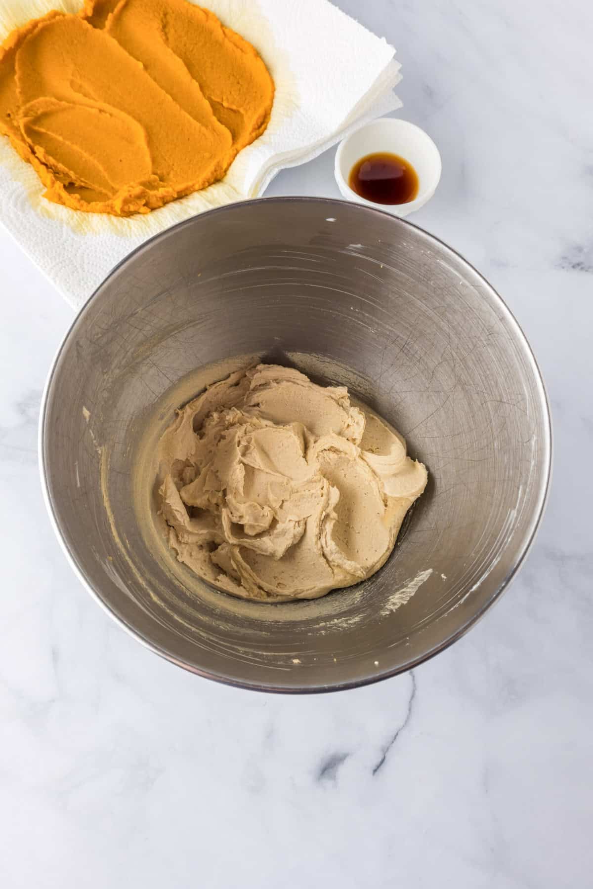 A metal mixing bowl with creamed butter and sugar in a bowl to make pumpkin chocolate chip bars with a plate with pumpkin puree on paper towels and a small bowl of vanilla nearby.