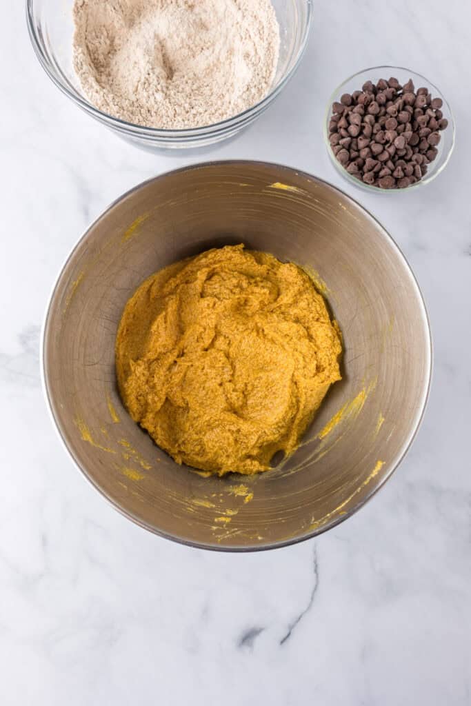 A metal bowl with orange cookie dough from mixed wet ingredients for pumpkin chocolate chip bars, with bowls of dry ingredients and chocolate chips nearby.