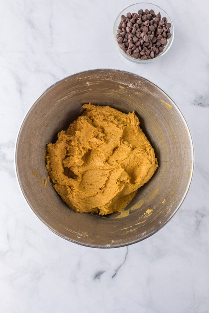 A mixing bowl filled with cookie dough for pumpkin chocolate chip bars with a bowl of chocolate chips nearby.