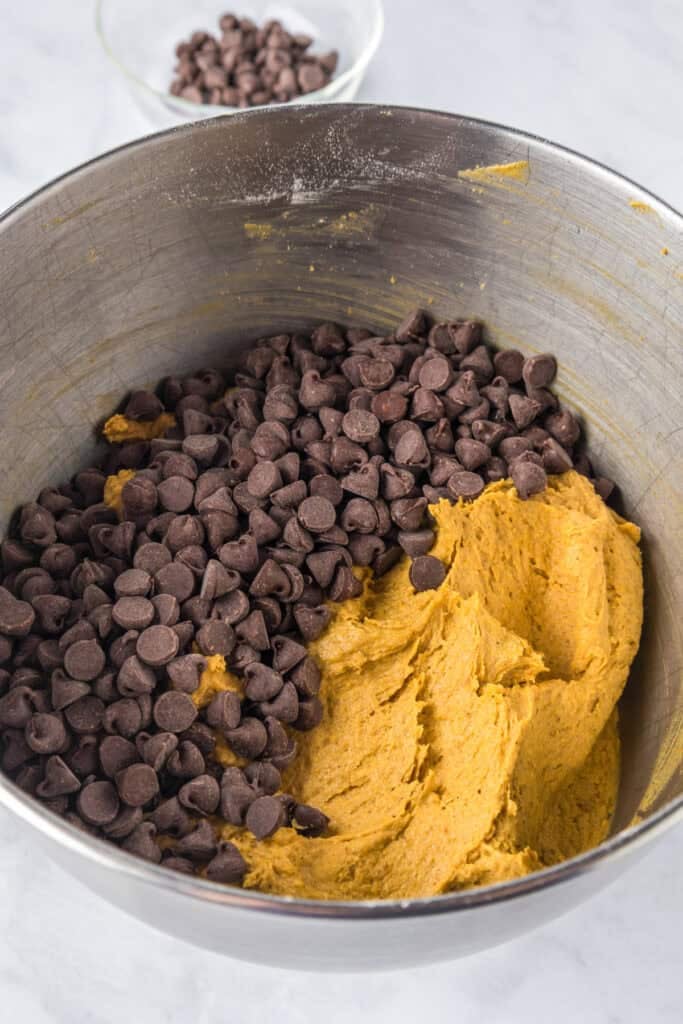 A metal mixing bowl full of cookie dough for pumpkin chocolate chip bars with chocolate chips being mixed into the dough.