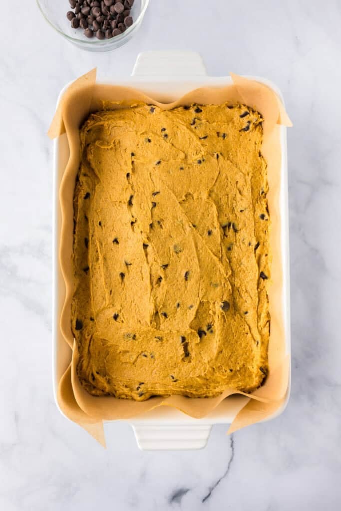 A parchment-lined baking dish filled with raw pumpkin chocolate chip bars cookie dough on a counter.