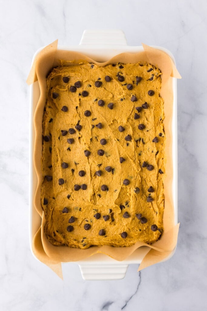 A parchment-lined baking dish filled with raw pumpkin chocolate chip bars cookie dough topped with chocolate chips sprinkled on top.