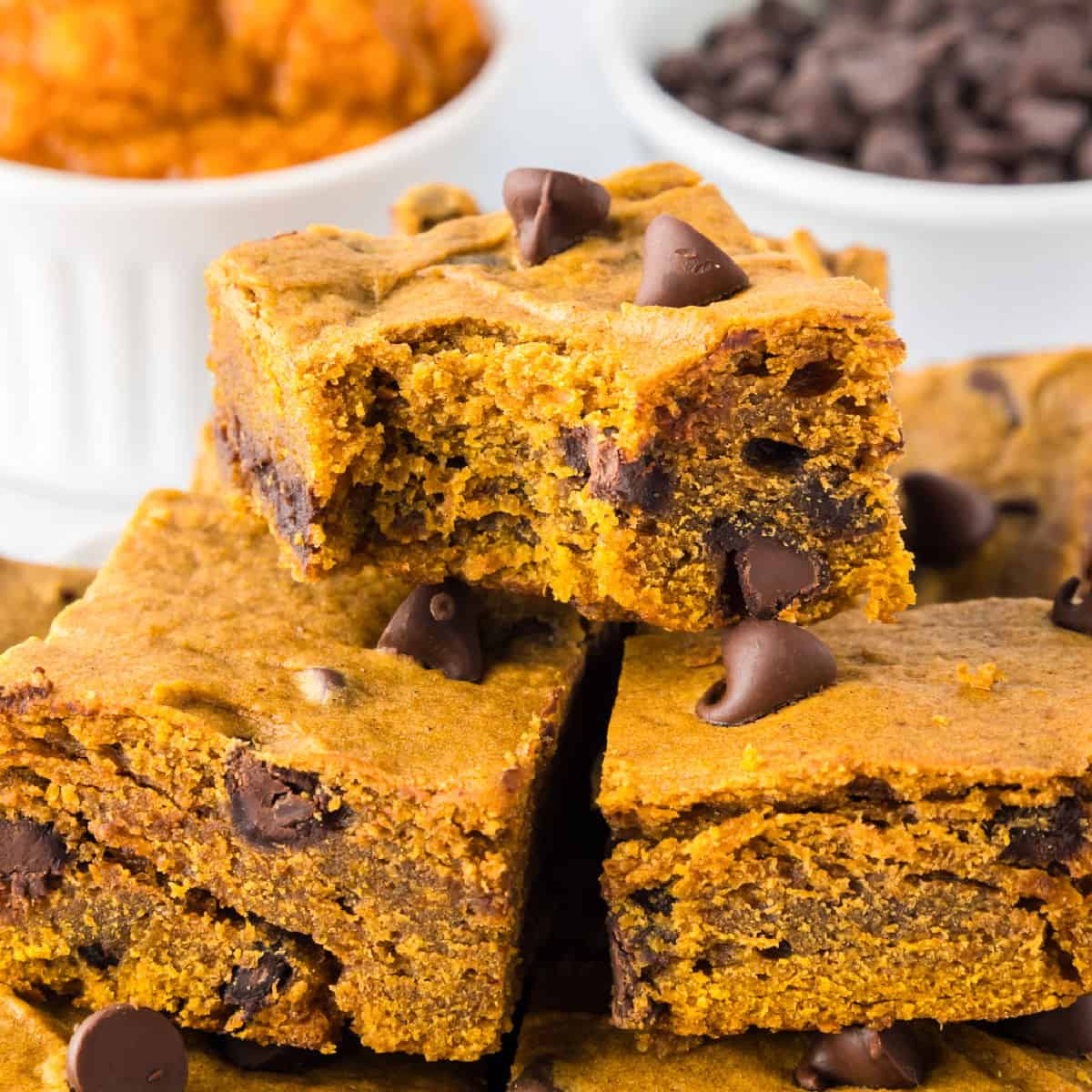 Three pumpkin chocolate chip bars are stacked on top of each other on a platter, with chocolate chips visible inside and a bite missing from the top bar.