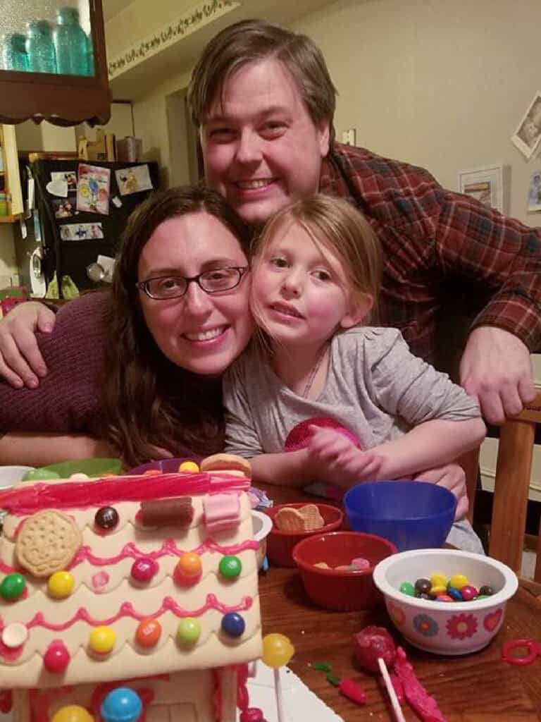 Three people, two adults and a child, pose and smile together at a table with a decorated gingerbread house and bowls of candy.