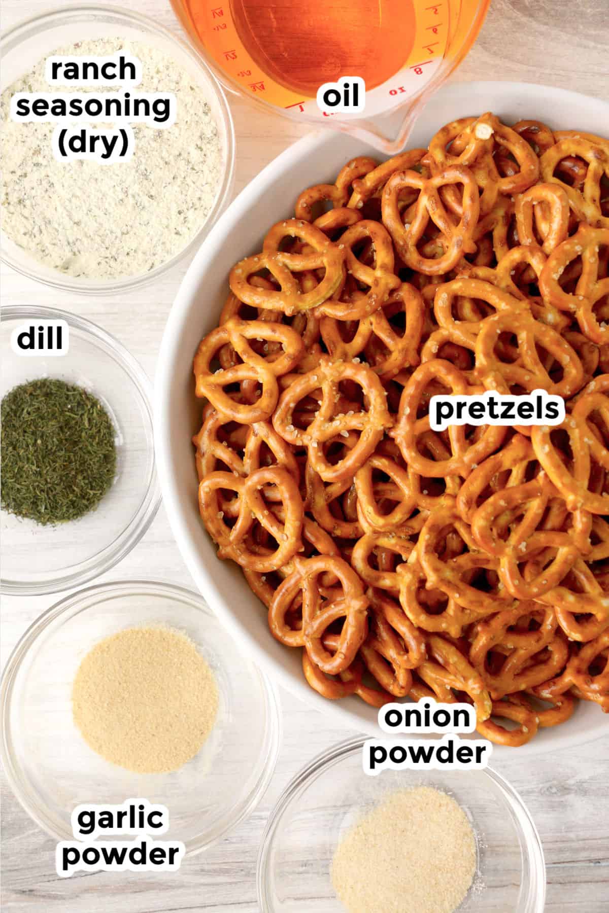 A bowl of pretzels is surrounded by small, labeled bowls filled with dry ranch seasoning, oil, dill, onion powder, and garlic powder to make garlic ranch pretzels.