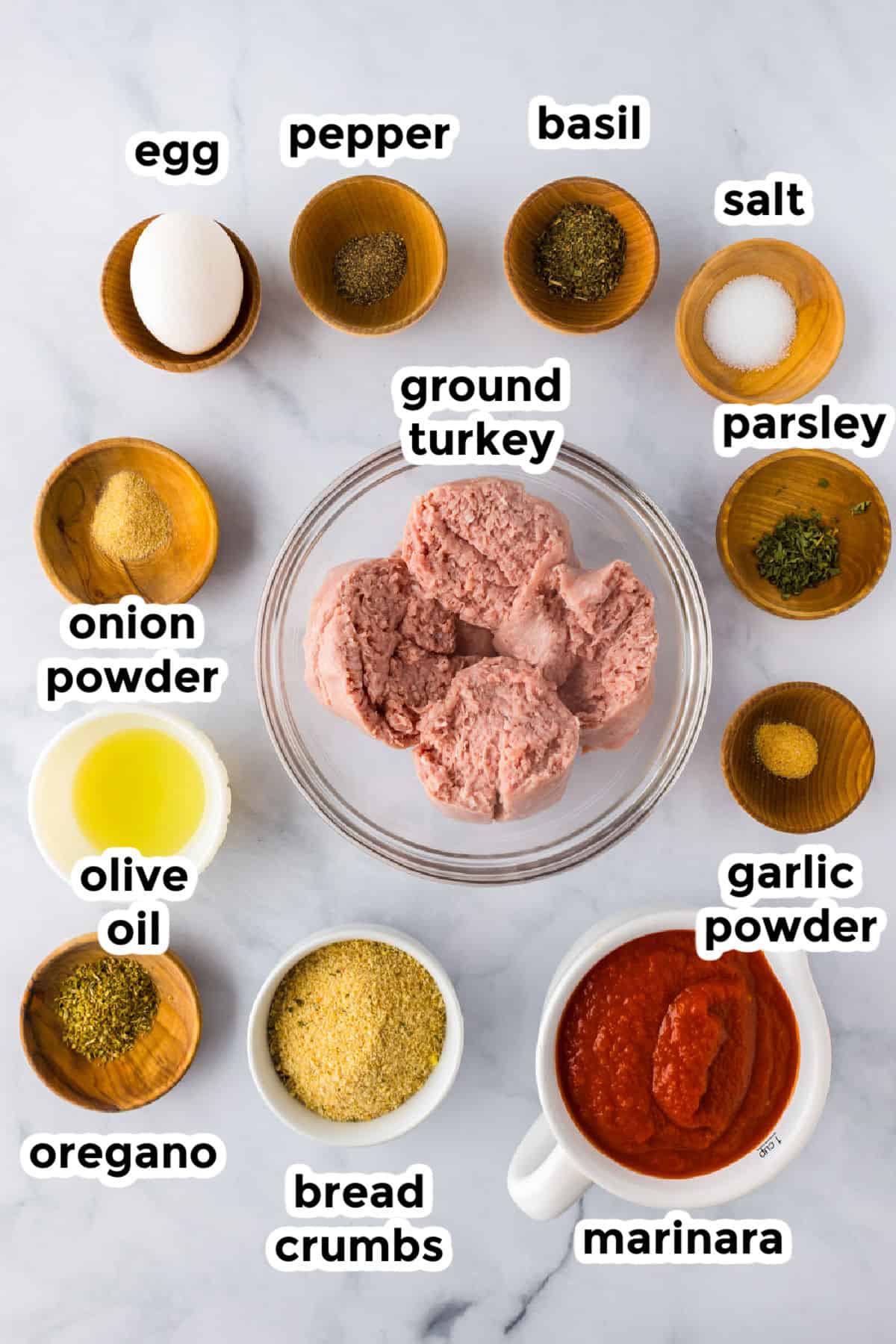 Ingredients for ground turkey meatballs are arranged in bowls on a marble surface: ground turkey, egg, marinara, bread crumbs, olive oil, and various spices including salt, pepper, and herbs.