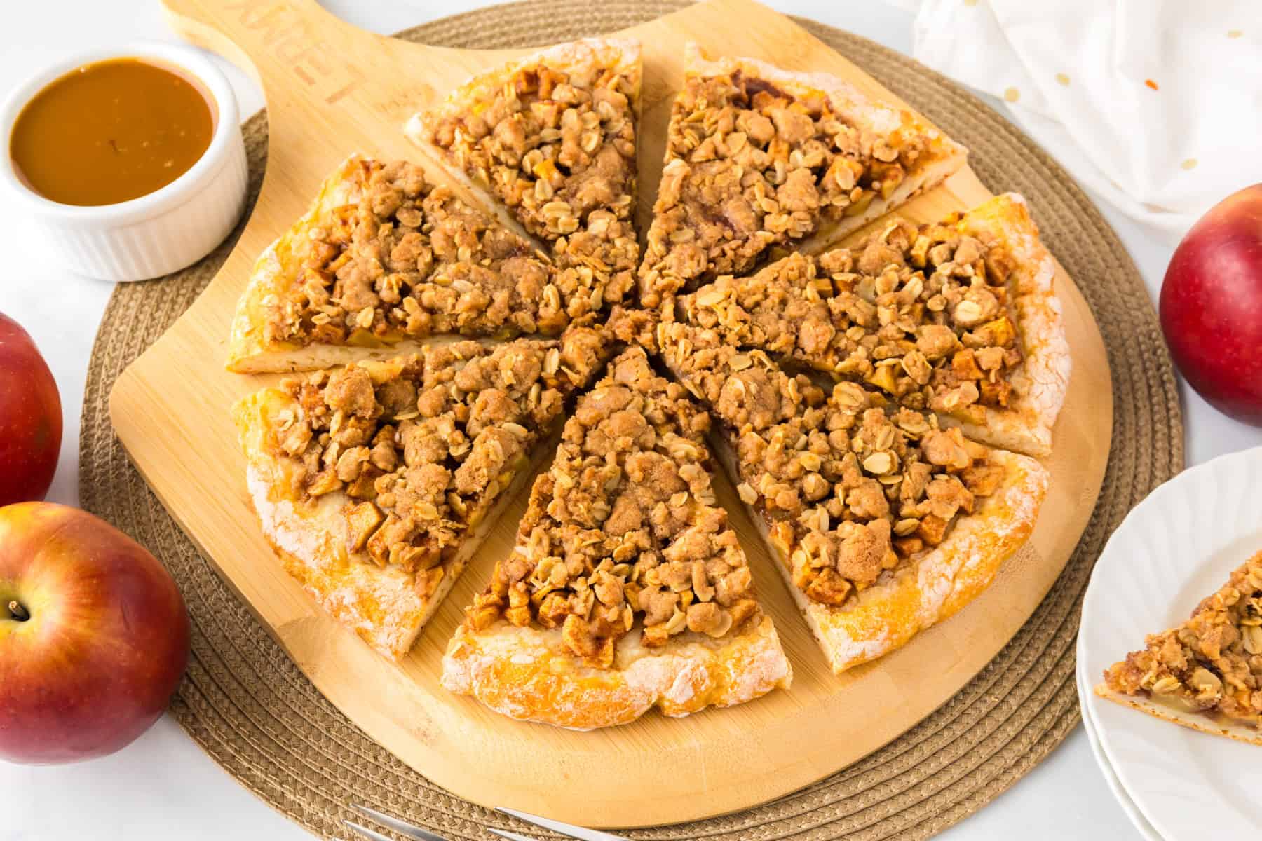 A sliced apple dessert pizza on a wooden board, surrounded by fresh apples, a bowl of caramel sauce, and a white plate.