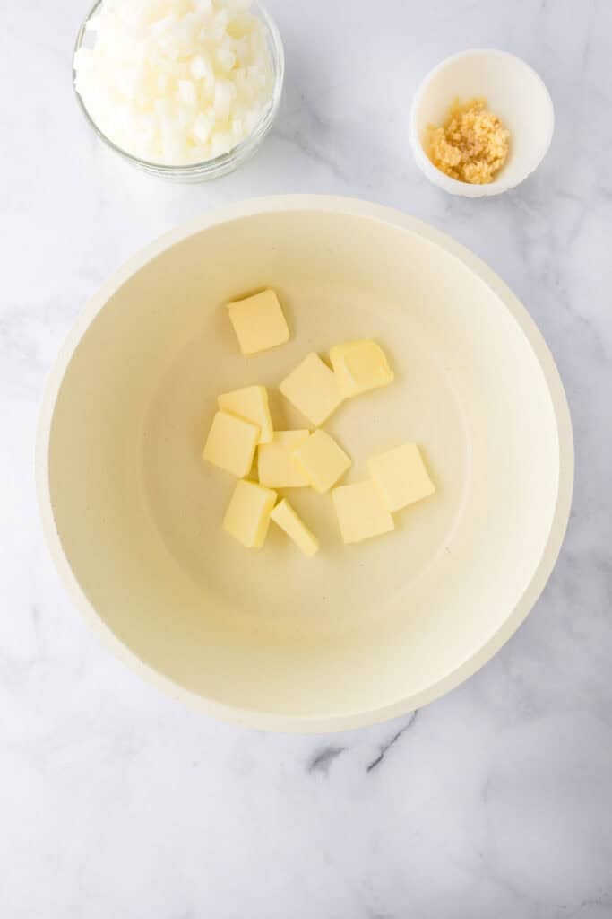 A bowl with cubed butter, next to a small bowl of minced garlic and a bowl of chopped onions—perfect prep for making a flavorful green bean casserole—on a marble surface.