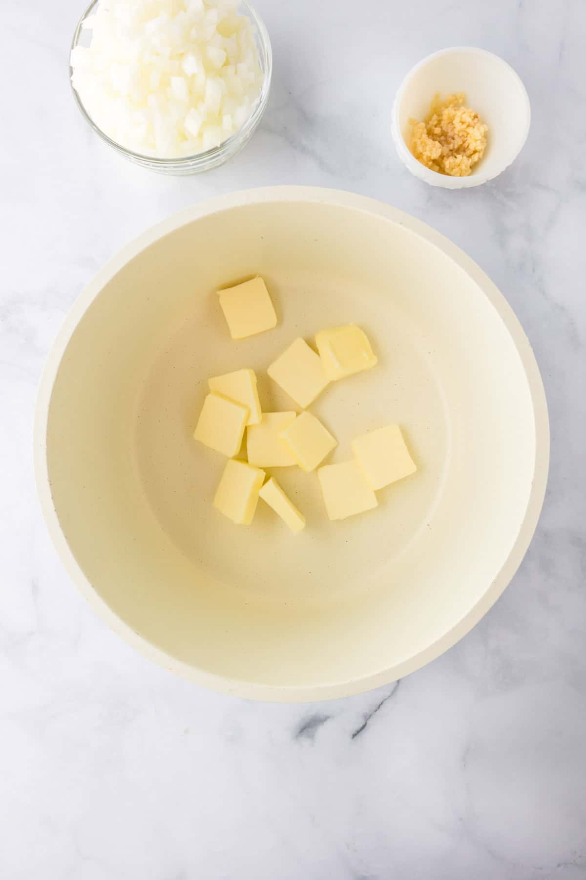 A bowl with cubed butter, next to a small bowl of minced garlic and a bowl of chopped onions—perfect prep for making a flavorful green bean casserole—on a marble surface.