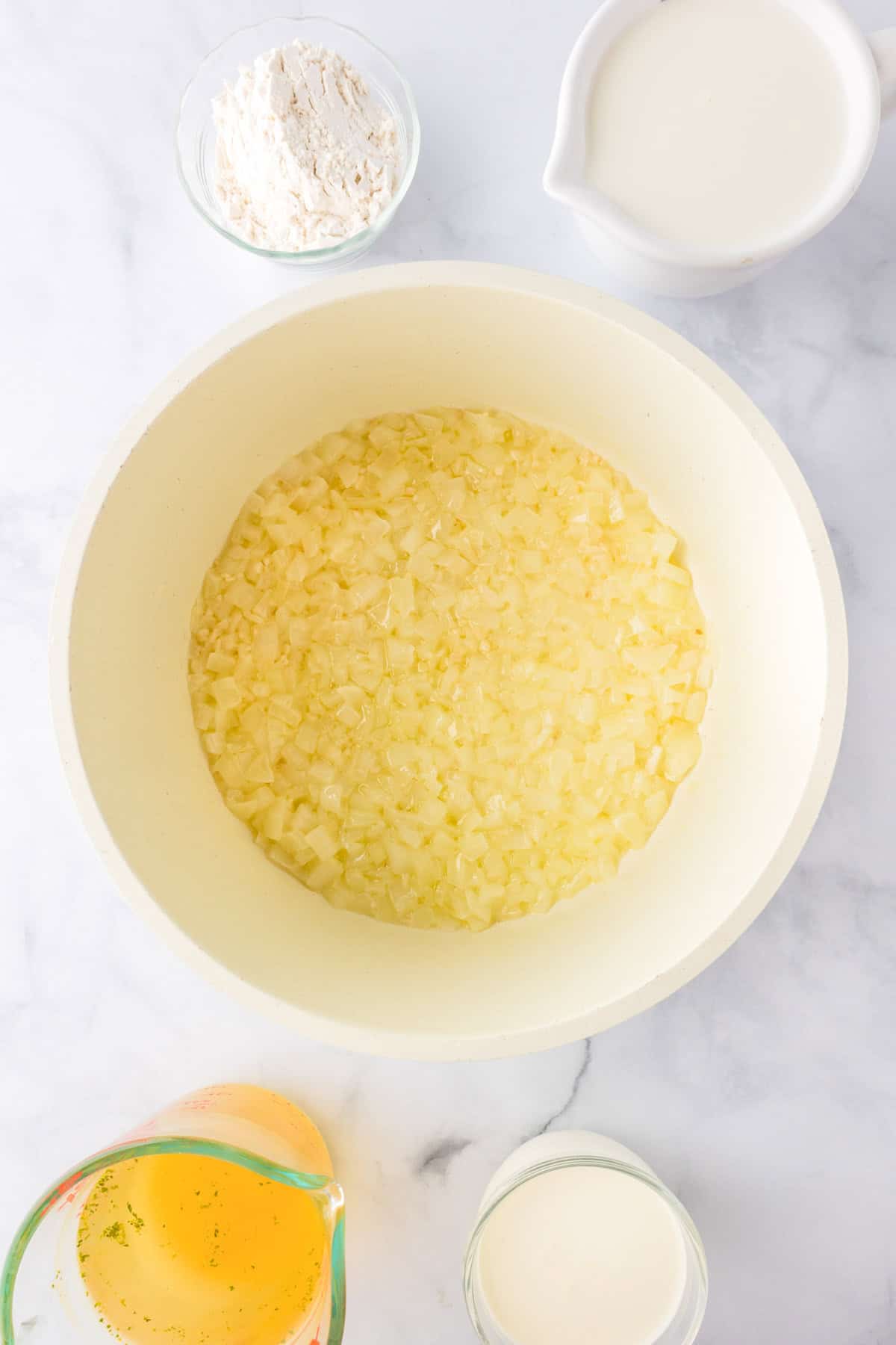 A bowl of diced onions sits in the center, surrounded by small bowls of flour, milk, cream, and a measuring cup of broth—key ingredients for preparing a classic green bean casserole—on a white surface.